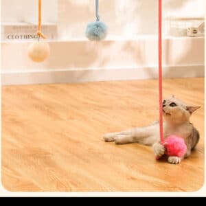 Playful gray and cream cat grips a pink pom-pom toy on a warm wooden floor.