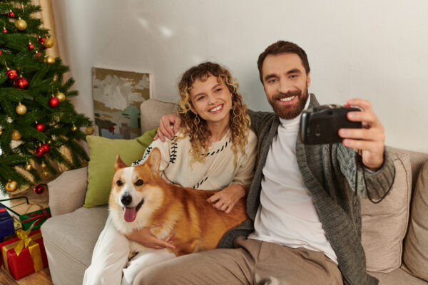 Joyful couple with corgi by Christmas tree, capturing festive moments together.